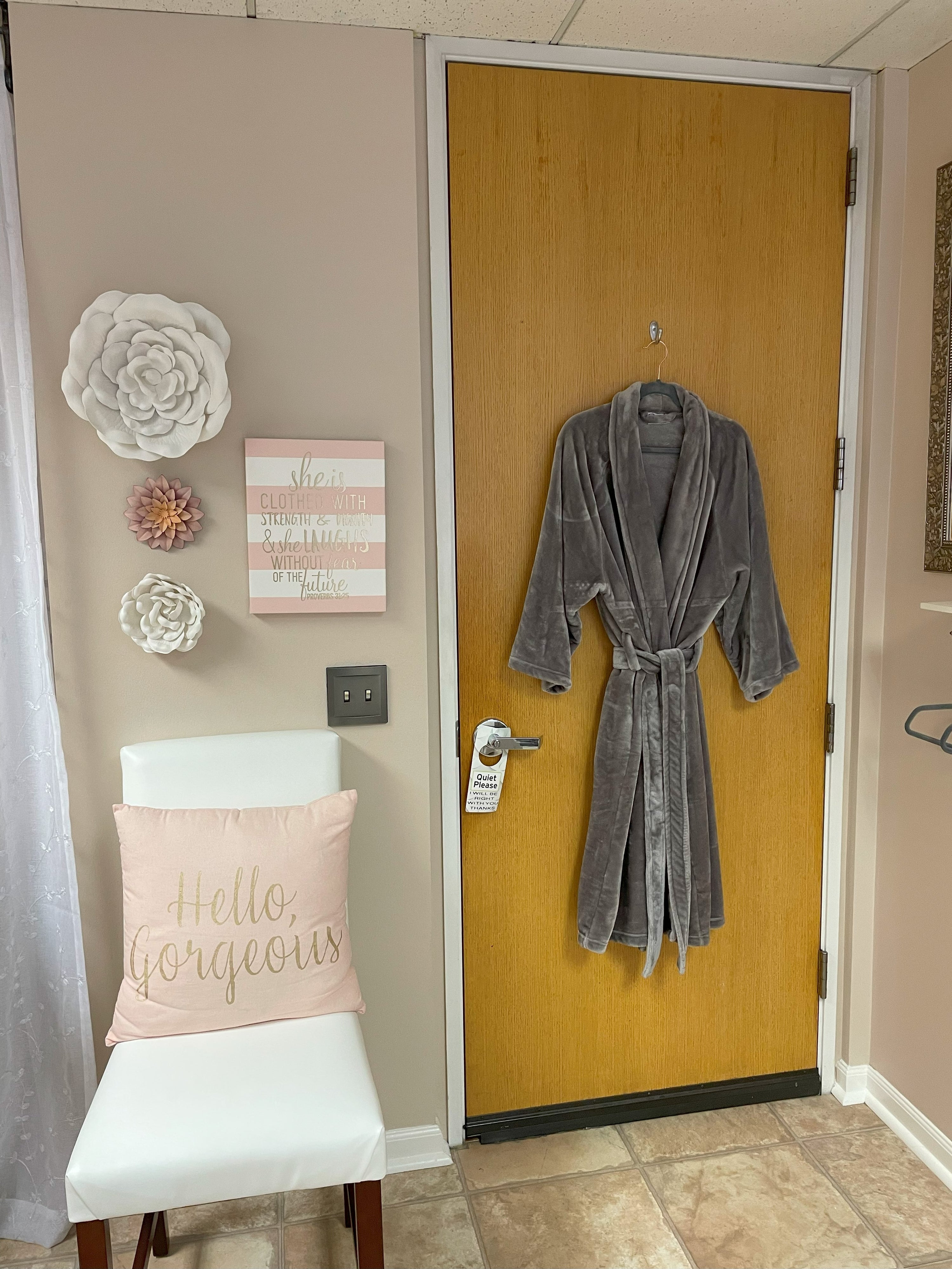 White chair with pink Hello Gorgeous pillow, pink and white picture with Proverbs 31:25 She is clothed with strength and dignity and she laughs without fear of the future, and plush and soft gray robe hung on door