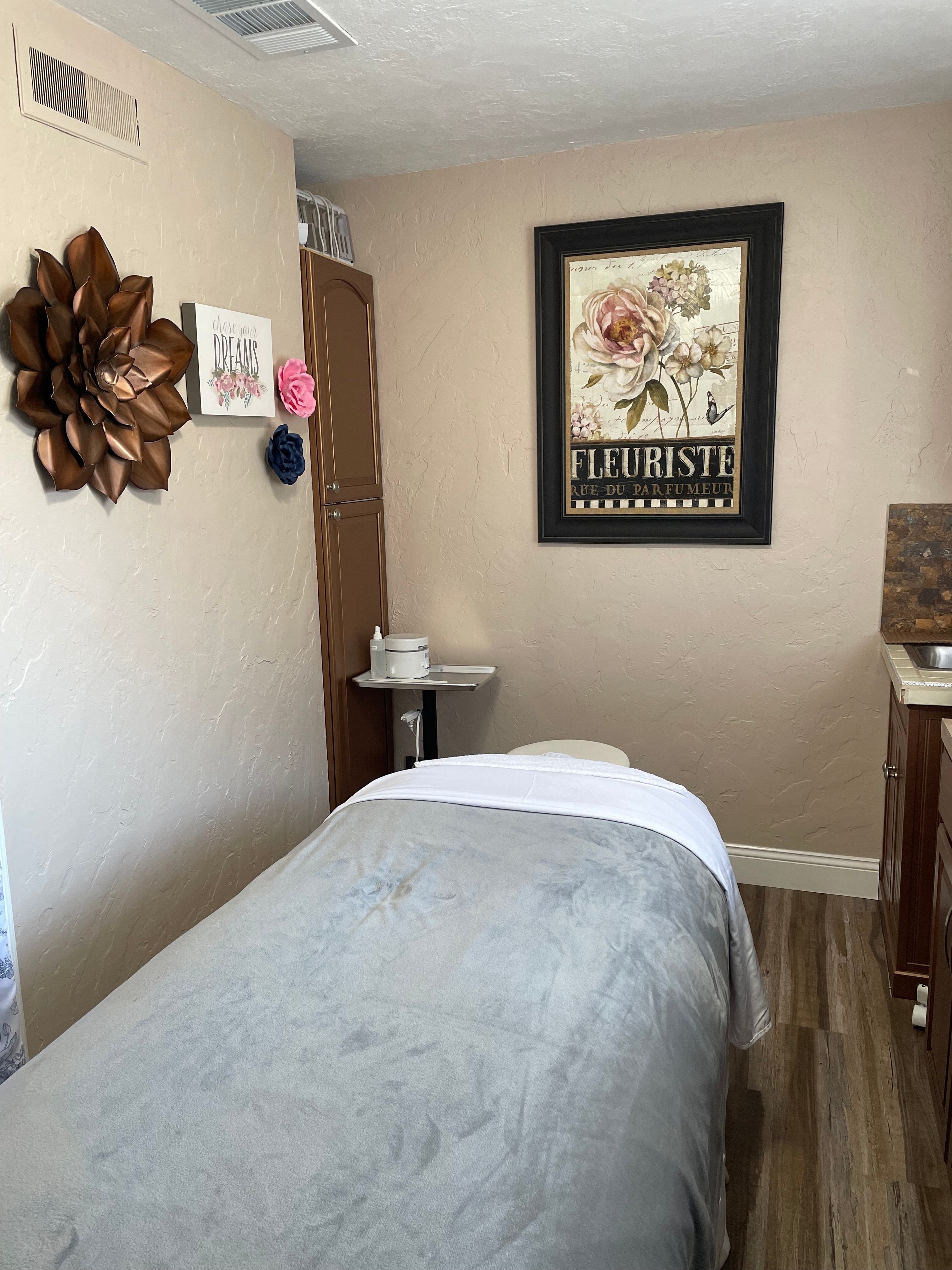 A serene treatment room with beautiful pink and copper floral pictures.  The facial bed is covered with a gray soft and plush blanket for a relaxing facial treatment.